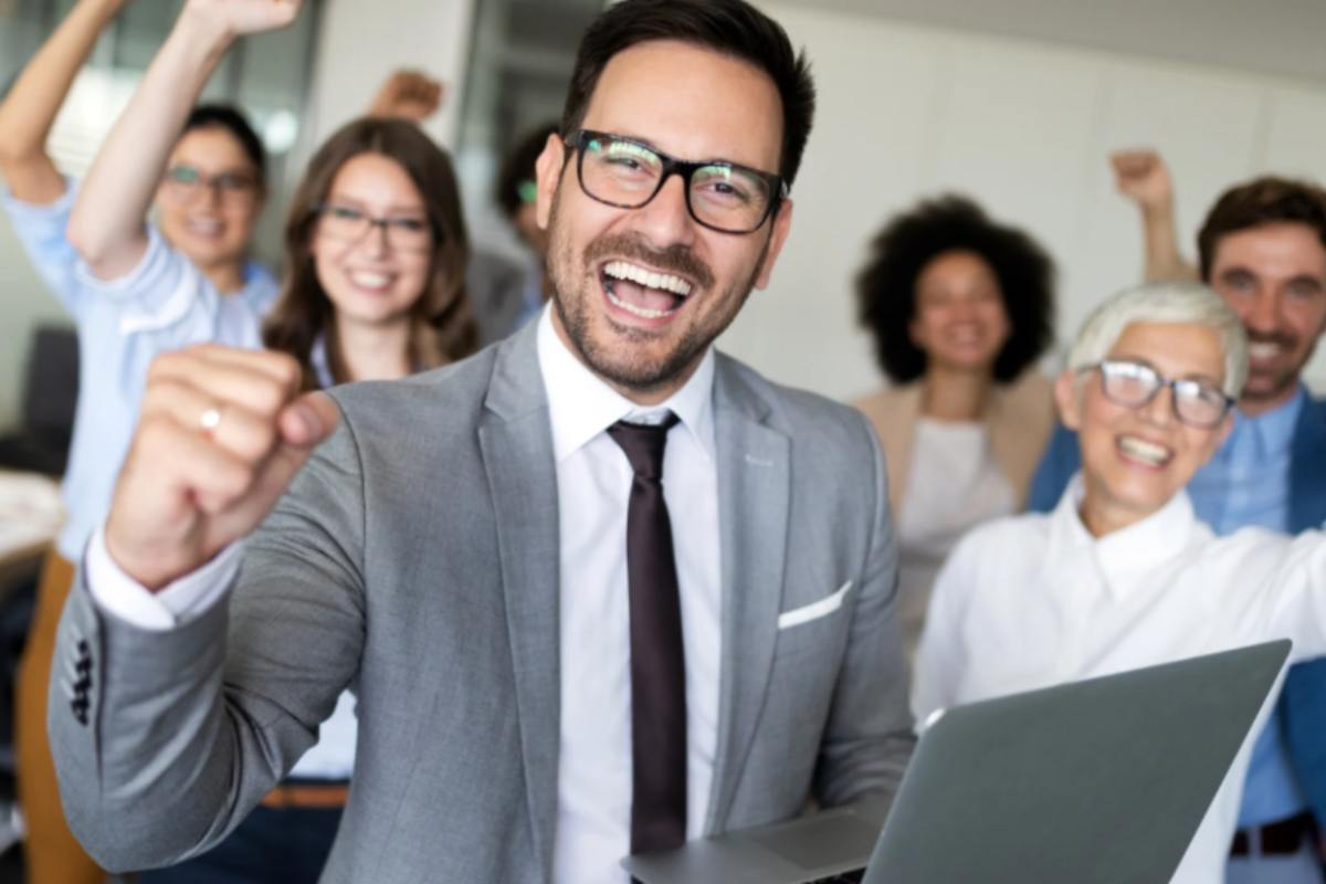 uomo con un computer in mano che esulta e dietro di lui persone che sorridono